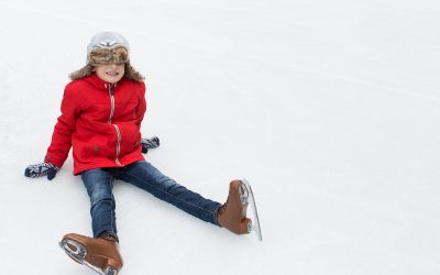 positive happy boy being silly and sitting on ice enjoying winter vacation at outdoor ice skating rink